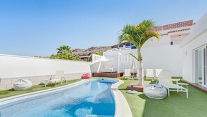 Un espace piscine extérieur moderne, doté d’une piscine aux lignes courbes, de chaises longues blanches et d’un palmier.