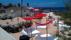 Un complexe hôtelier en bord de mer, avec des chaises longues blanches, des parasols rouges et un petit bâtiment.