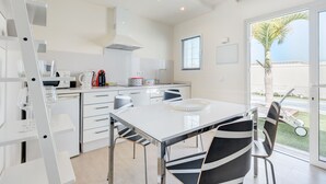 Une cuisine moderne avec des meubles blancs, une table à manger et des chaises. On y trouve une fenêtre avec des stores, une vue sur un palmier à l’extérieur et un balcon avec des meubles d’extérieur.