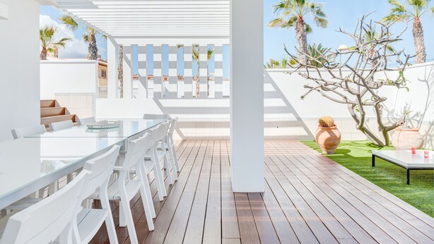 Une terrasse extérieure moderne, dotée d’une table en verre et de chaises blanches, d’une terrasse en bois et offrant une vue sur des palmiers et un ciel dégagé.