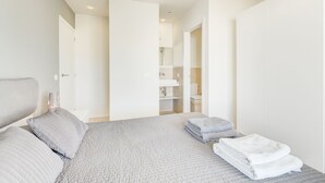 Une chambre à coucher moderne avec un grand lit, une table de chevet et une salle de bain visible à travers une porte ouverte.