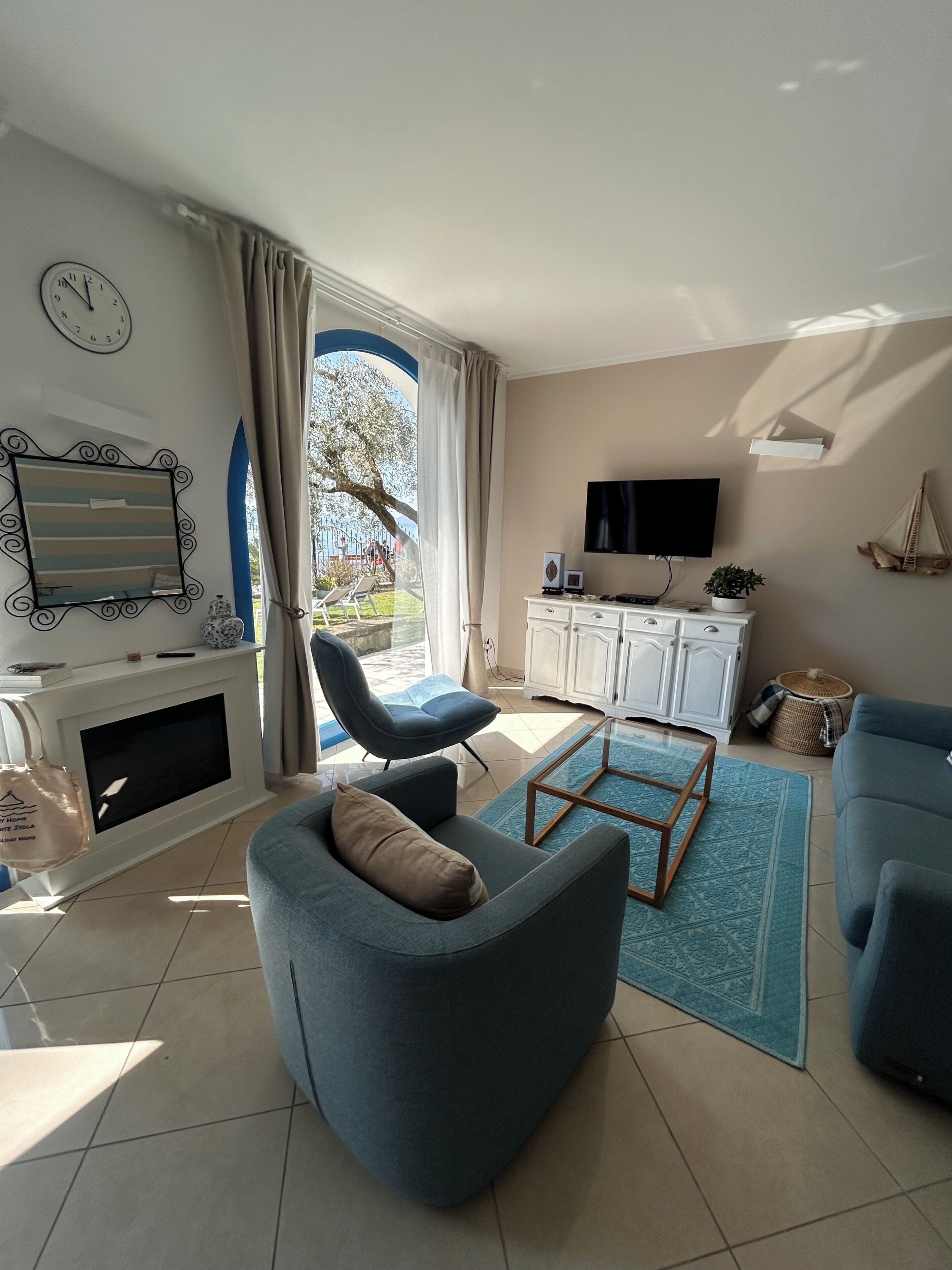 A modern living room with a sofa, armchair, and a coffee table. There is a television mounted on the wall, a fireplace, and a window with curtains.