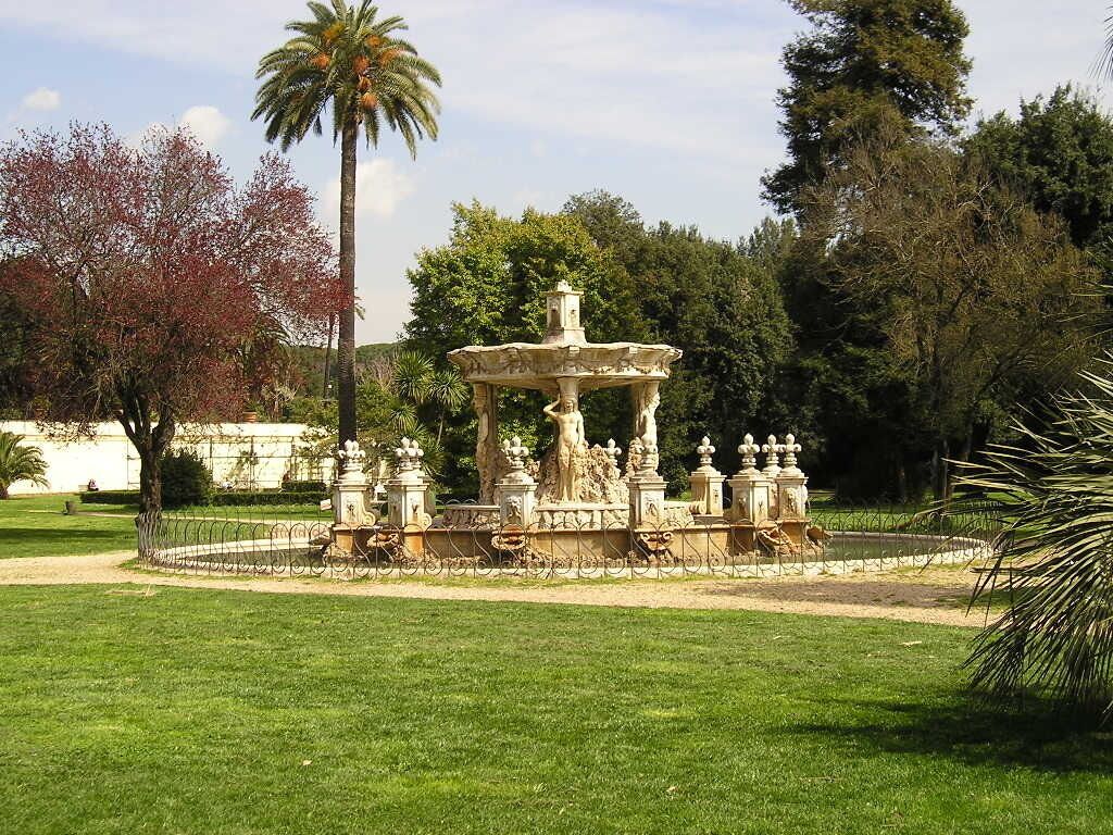 Una fuente con una cuenca central y varias estatuas, rodeada por una cerca y follaje.