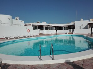 Une piscine à l’eau d’un bleu limpide, entourée de murs blancs et dotée d’un espace couvert avec des fenêtres.