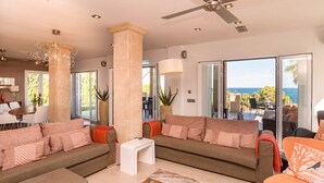Un salon spacieux avec deux canapés, une table basse et un ventilateur de plafond. De grandes fenêtres offrent une vue sur l’extérieur.