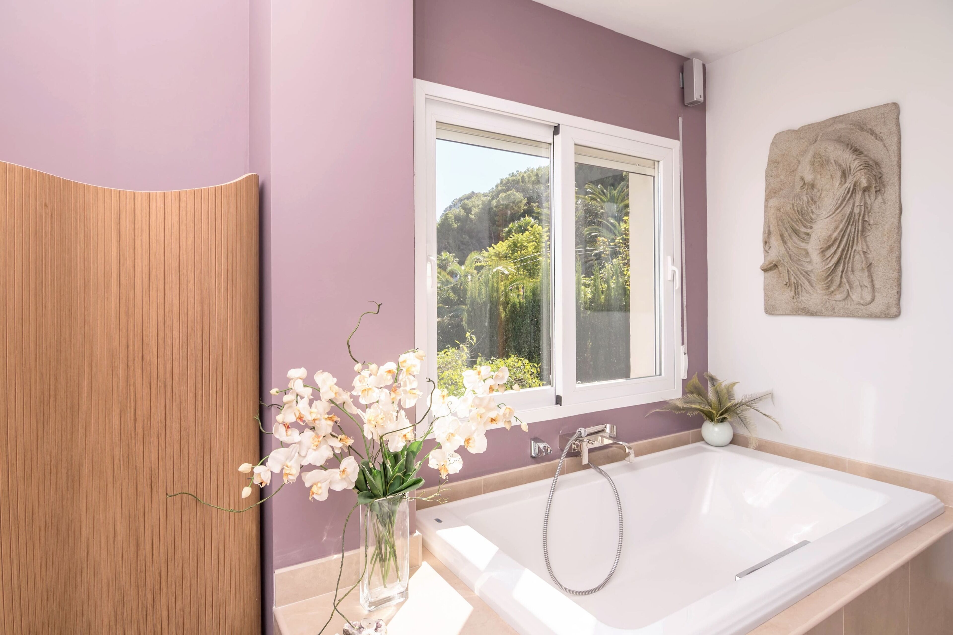 Une salle de bain avec une tête de lit en bois, une baignoire blanche, une fenêtre donnant sur un paysage verdoyant et une petite plante en pot.