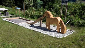 Una escultura de madera de un caballo en un jardín, con un área pavimentada y un cartelón de señalización al fondo.