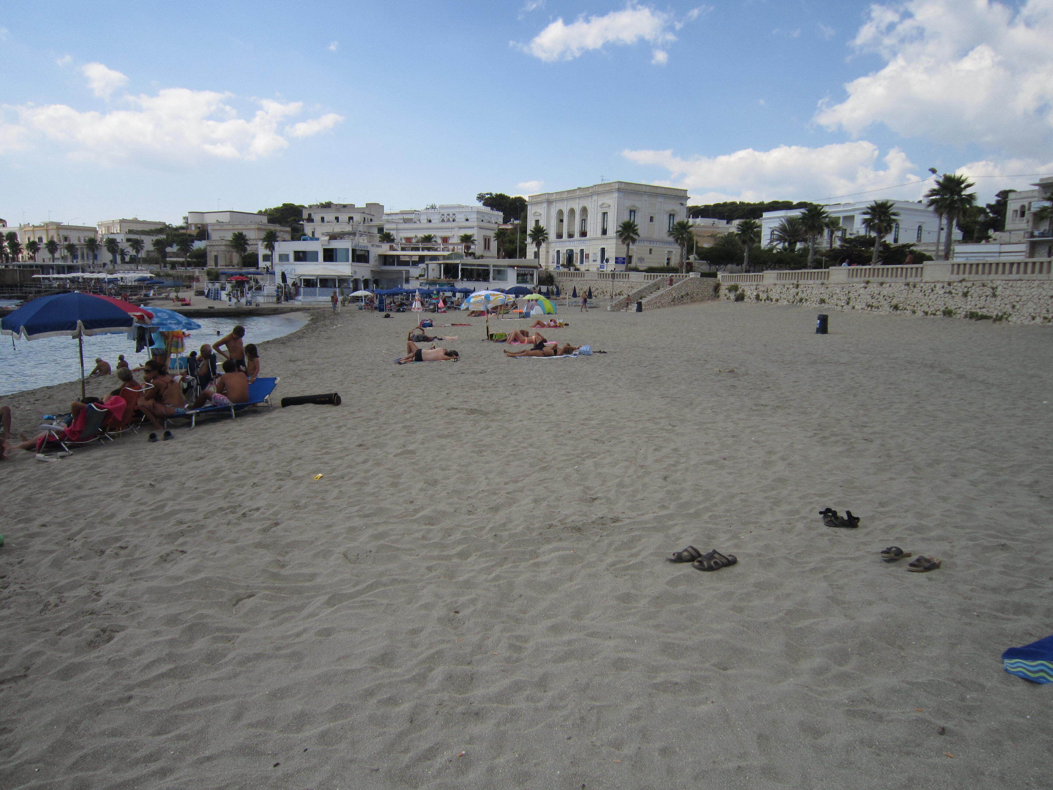 A beach with people sunbathing and swimming, umbrellas, and a building in the background.
