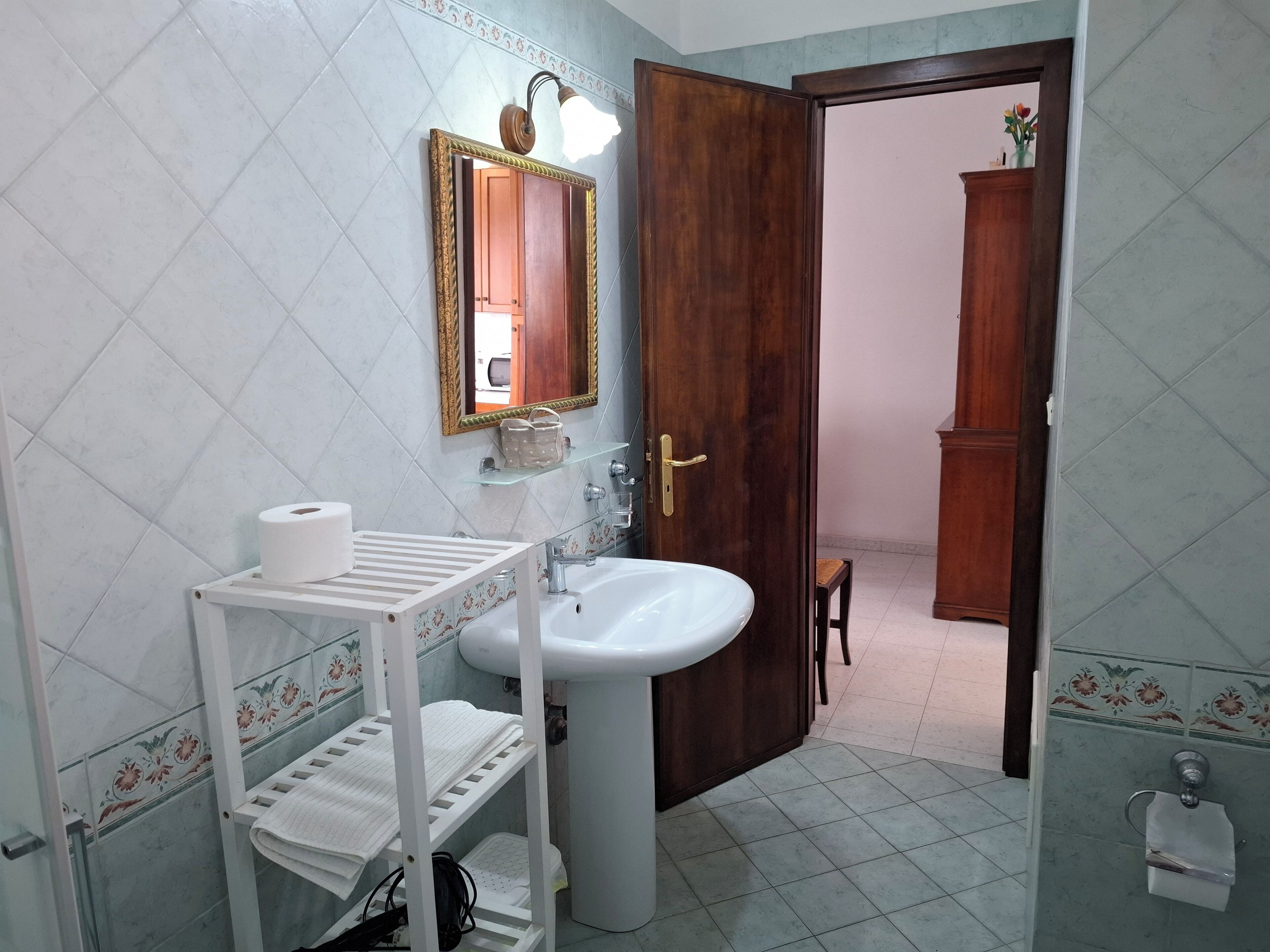 Un baño con lavamanos blanco, un espejo con luz, una puerta de madera y un estante con artículos de aseo.