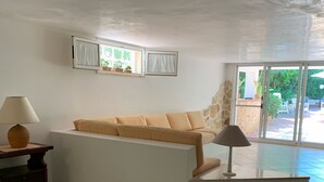 A room with a sofa, a dining table with chairs, a lamp, and a view of an outdoor area with plants and a pool. - Greek/Mediterranean style villa of 200 sqm, with elegant interiors, family friendly (Pilone)