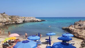 A beach with clear turquoise water, rocky cliffs, and several people swimming and sunbathing.
