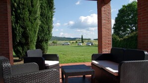 An outdoor patio with wicker furniture, a brick wall, and a view of a grassy field with parked cars and distant hills.