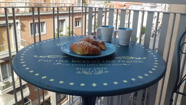 Un balcon avec une table sur laquelle se trouvent un croissant, deux tasses blanches et une assiette. La table porte un texte et un logo.