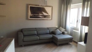 Un salon avec un canapé d’angle gris, un tableau encadré représentant un voilier accroché au mur, un radiateur et une fenêtre avec des rideaux.