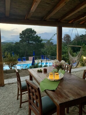 Ein Essbereich im Freien mit Holztisch, Stühlen und einer Hängematte. Im Hintergrund ein Pool, umgeben von blauen Liegestühlen und Blick auf Bäume und Berge.