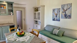 Un salon chaleureux avec un canapé vert, une table à manger agrémentée d’un panier de fruits et un coin cuisine avec un meuble et une porte décorative.