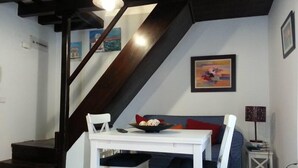 Un área de comedor con una mesa y sillas blancas, un sofá azul con cojines cuadriculados, una escalera y un cuadro en la pared.