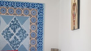 Una cama bien hecha con colcha blanca, almohadas rojas y una toalla blanca doblada. Sobre la cama, un panel decorativo con diseños azules y anaranjados.