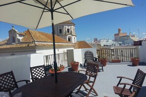 Een terras op het dak met een tafel, stoelen, plantenbakken en een uitzicht op een kerk met een klokkentoren.