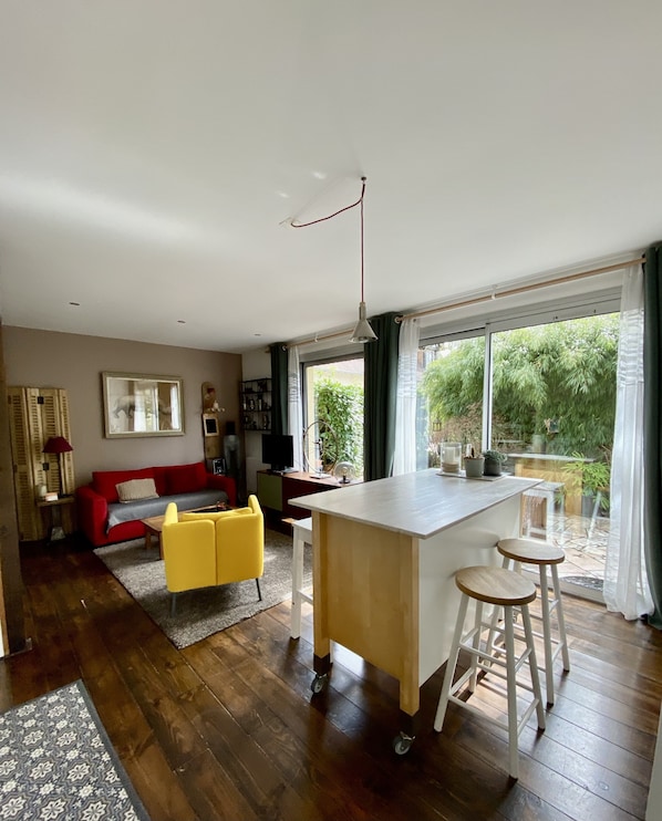 A modern living room with a red sofa, yellow chairs, a wooden dining table with stools, and a large window with a view of greenery.