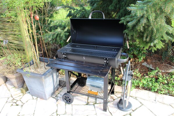 A portable barbecue grill with a metal frame and wheels, placed on a stone patio surrounded by greenery.