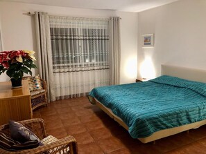 Ein Schlafzimmer mit Bett, Nachttisch, Stuhl, Tisch mit Pflanze und Fenster mit Vorhängen.
