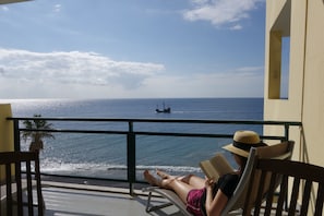 Varanda com vista para o mar, uma pessoa lendo um livro e um navio ao longe.