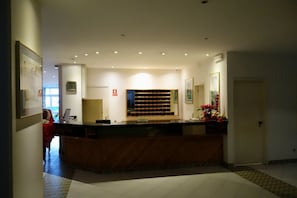 Ein Empfangsbereich mit einem hölzernen Empfangstresen, einem großen Fenster mit Gitterstruktur und einem roten Brandmelder an der Wand.