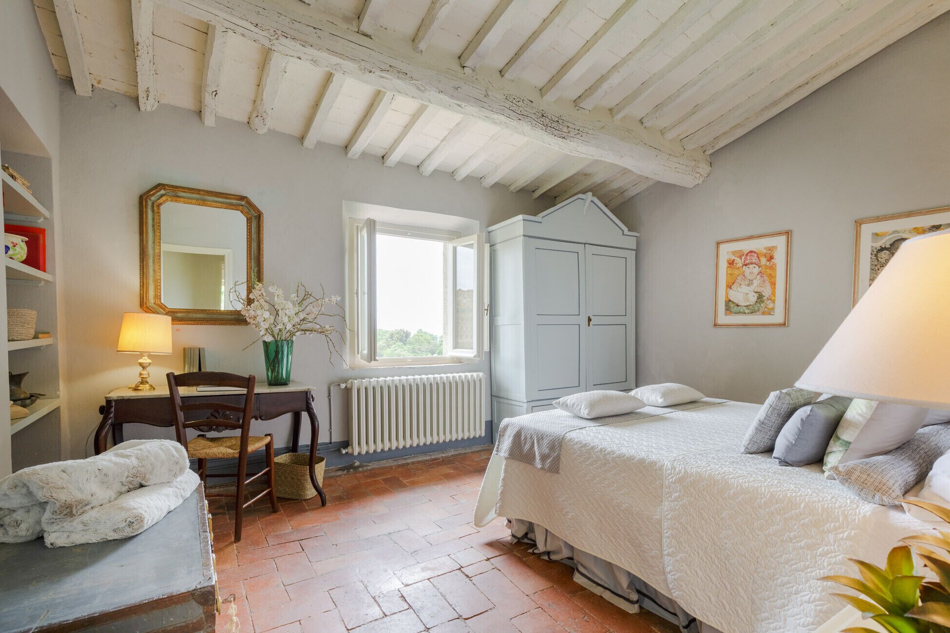 A bedroom with a bed, a chair, a table, a radiator, a wardrobe, and a window.