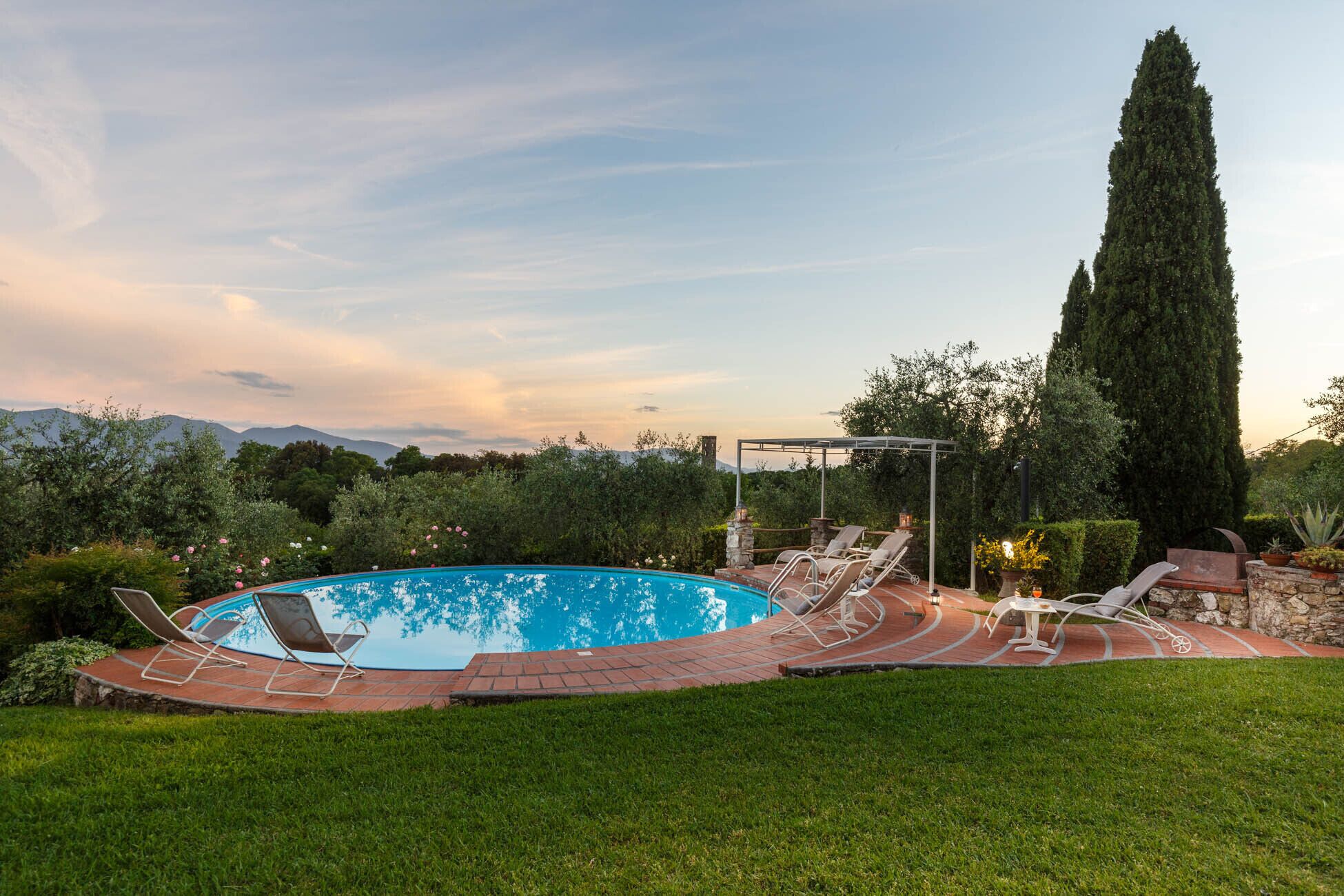 Une piscine avec des chaises longues, une pergola et un grand arbre dans un jardin aménagé.