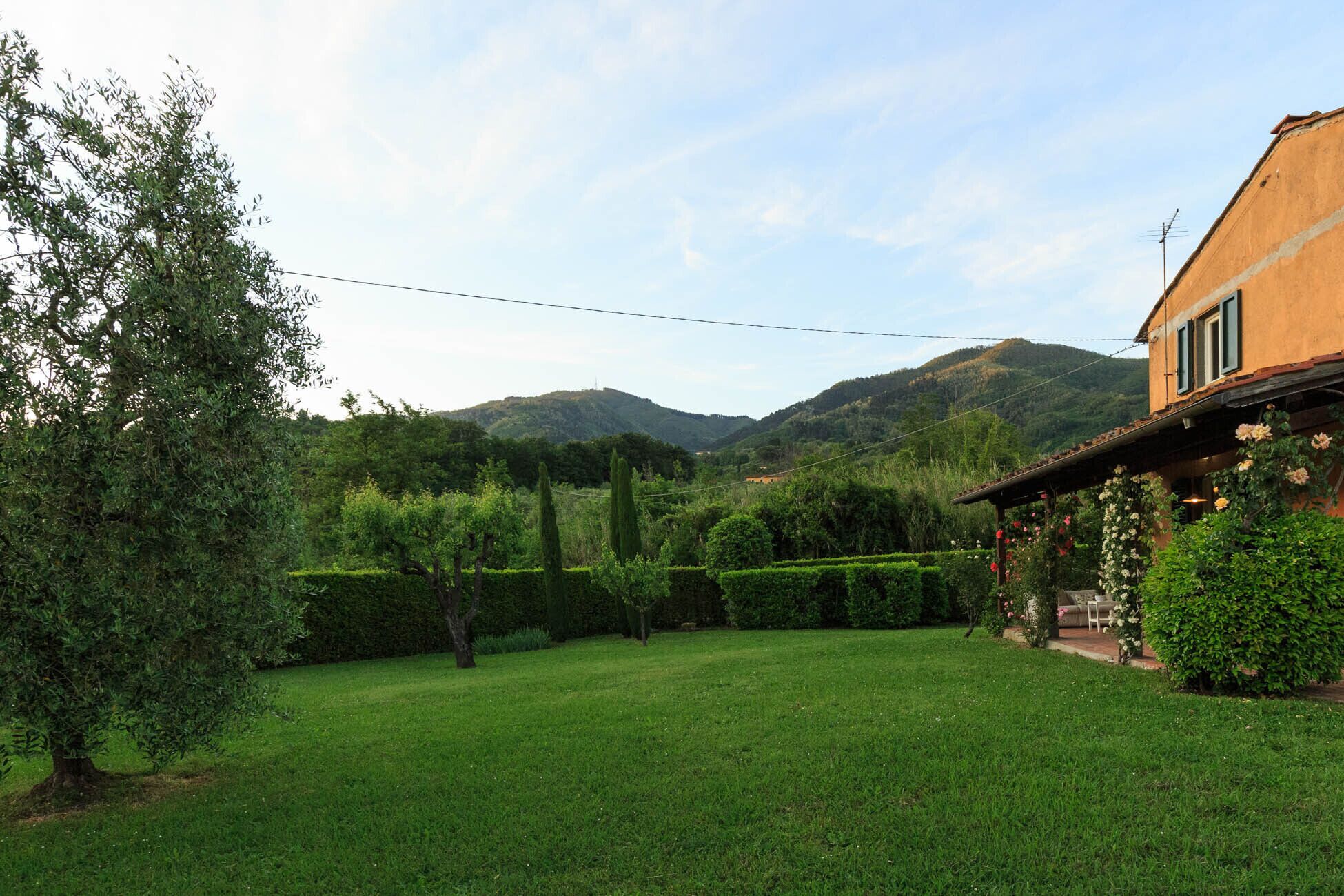A house with a well-maintained lawn, a pergola with climbing plants, and a mountainous backdrop.