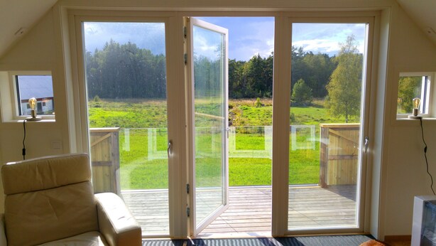 Una habitación con puerta corrediza de vidrio que da a una terraza de madera con vista a un campo cubierto de césped y árboles.