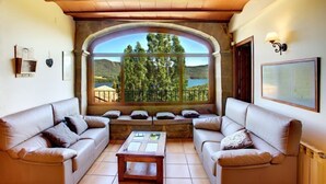 Una sala de estar acogedora con dos sofás y una mesa de centro, un amplio ventanal de arco que ofrece una vista panorámica de árboles y colinas, y techo de madera.
