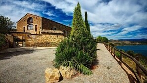 Edificio de piedra con fachada rústica, camino de grava y valla de madera que da a una masa de agua.
