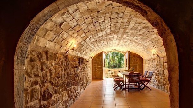 Un interior de piedra con arco, con una mesa y sillas, una ventana con vistas a la vegetación, y una pared de piedra.