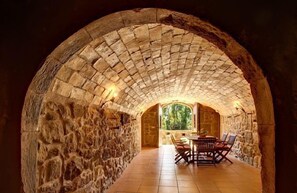 Interior arqueado de pedra com uma mesa e cadeiras, uma janela com vista para vegetação e uma parede de pedra.