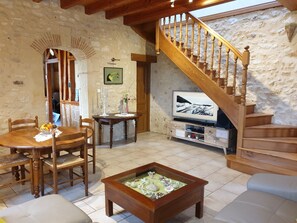 Un salon chaleureux avec un escalier en bois, une table à manger avec des chaises, une table basse agrémentée d’objets décoratifs et un téléviseur posé sur un meuble en bois.