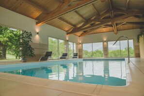 Una piscina interior con agua azul cristalina, vigas de techo de madera y grandes ventanales con vistas a zonas verdes.