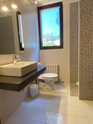 A modern bathroom with a white sink, mirror, and toilet. There is a window providing natural light and a view of greenery outside.