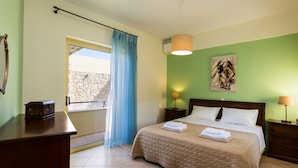 A bedroom with a bed, a nightstand, a lamp, a painting, a window with curtains, and a view of a stone wall. - Malena Villa with Private Pool | Walking distance to beach & restaurants (Rethymno)