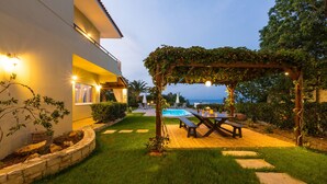 A well-lit outdoor area with a pool, a pergola with vines, and a wooden deck with a table and chairs. - Malena Villa with Private Pool | Walking distance to beach & restaurants (Rethymno)