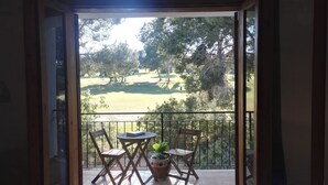 Un balcón abierto con vistas a un campo de golf, dos sillas de madera, una mesita y una planta en maceta.