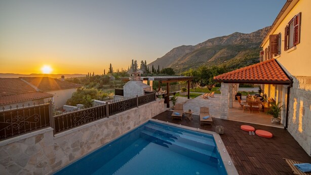 Une piscine bordée d’un mur en pierre et dotée d’une terrasse en bois, un espace de détente extérieur abrité et une vue sur les montagnes au coucher du soleil.