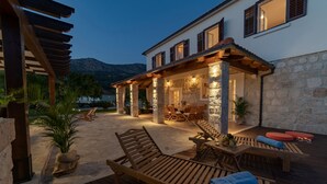 Une terrasse en bois avec des chaises longues et une table, un mur en pierre et un bâtiment doté d’une façade en pierre et de fenêtres.
