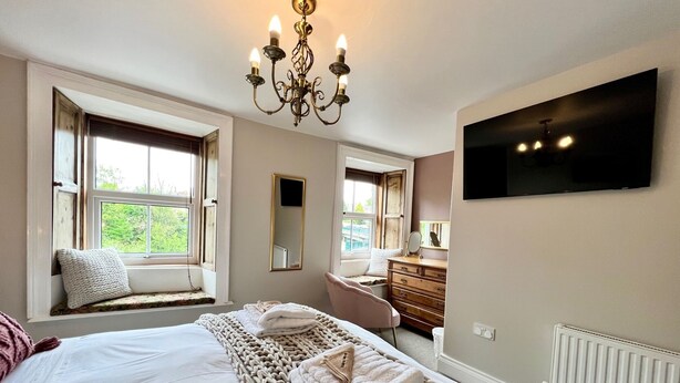 A bedroom with a large bed, a window seat, a dresser, a mirror, and a chandelier. - Luxury character Cottage in a popular Village location in Central Northumberland (Morpeth)