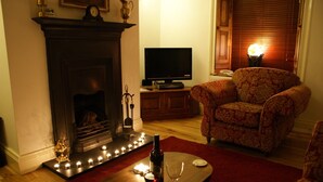 A living room with a fireplace, a television, a patterned armchair, and a table with a wine bottle and glasses. - Luxury character Cottage in a popular Village location in Central Northumberland (Morpeth)