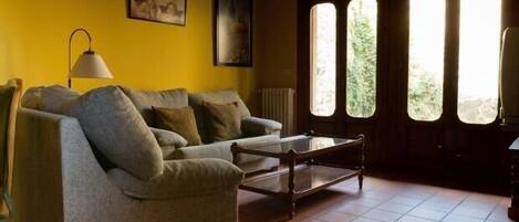 A living room with a sofa, a coffee table, and a lamp. There are two windows with a view of greenery outside.
