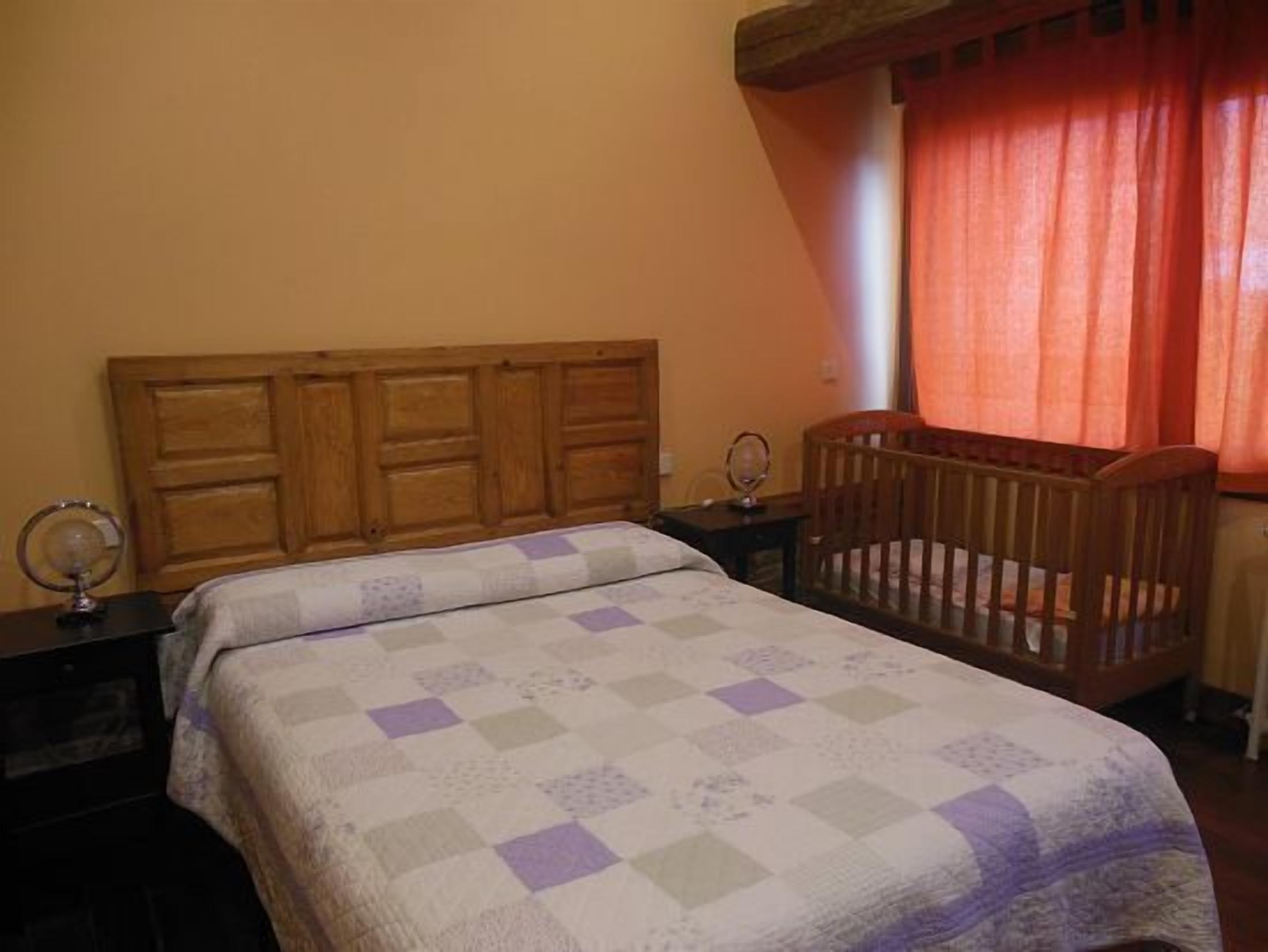 A bedroom with a wooden headboard, a crib, a bedside table with a mirror, and a window with curtains.