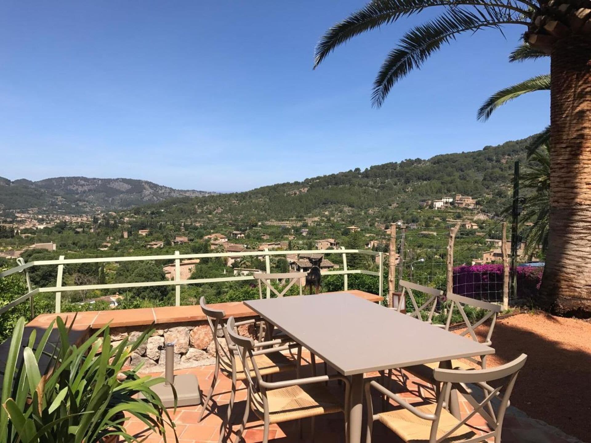 Eine Terrasse im Freien mit Tisch und Stühlen, mit Blick auf eine hügelige Landschaft mit Gebäuden.