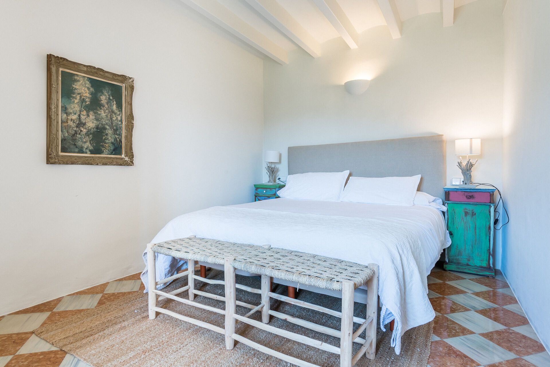 Une chambre à coucher avec un lit, un banc, une table de chevet, une lampe et un tableau au mur.
